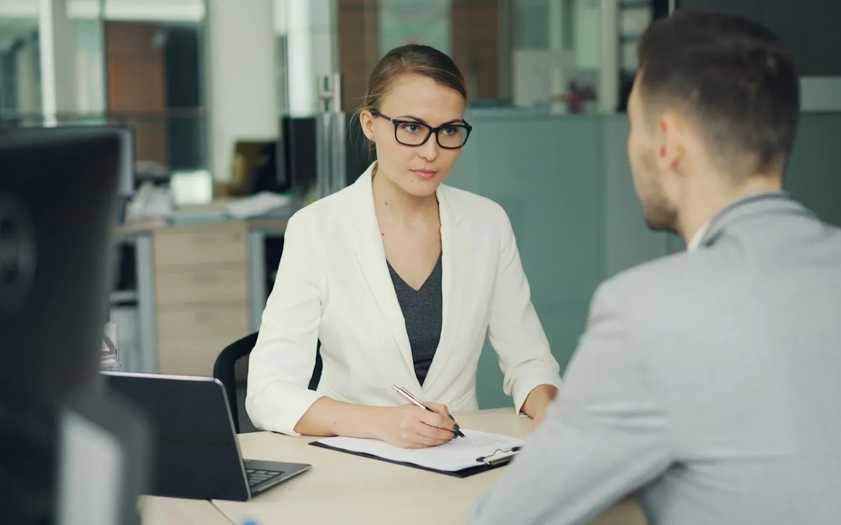 A manager taking notes while interviewing an employee across a desk, one of the main training needs analysis methods