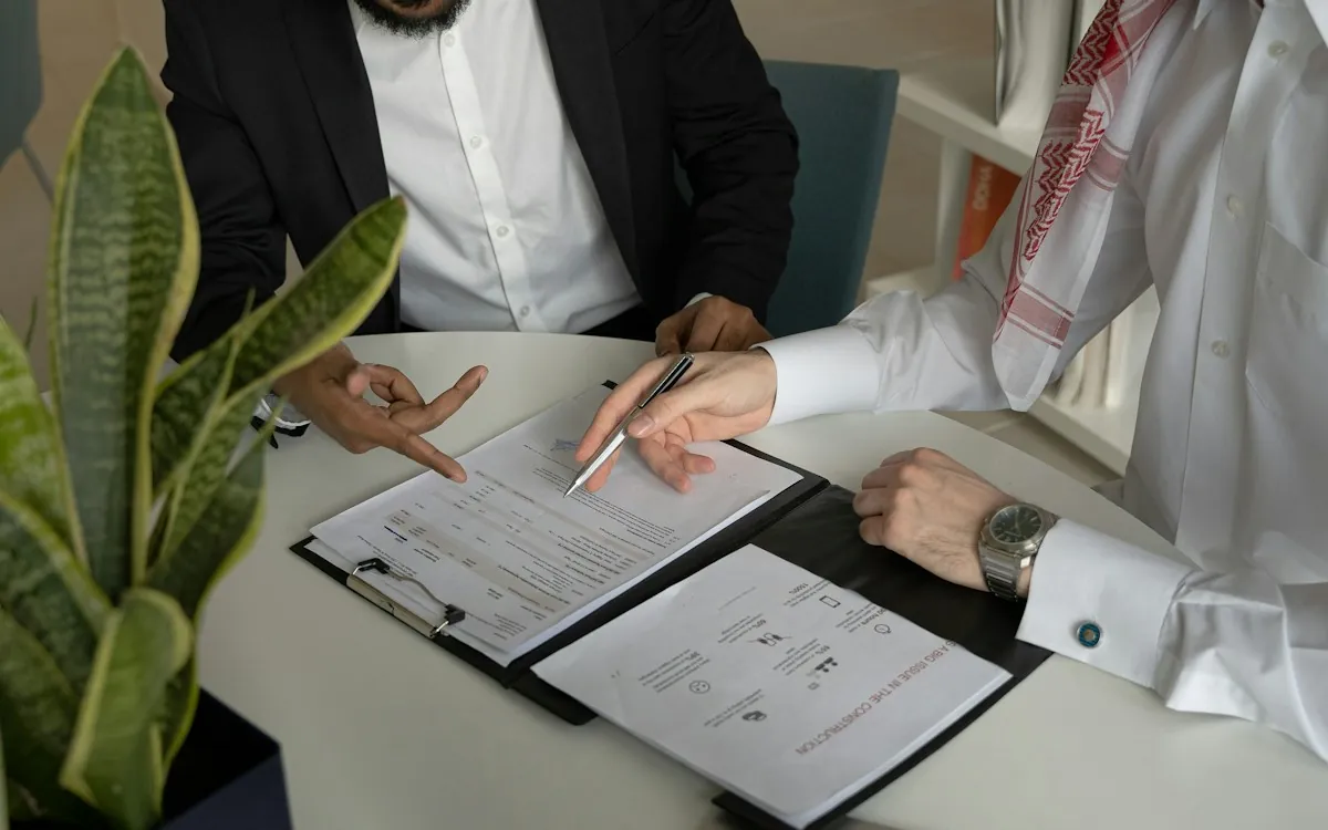 Two business partners at a table going through the clauses of a printed outsourcing contract with a pen